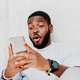 Young african american man with surprised facial expression hold smartphone in his hands, typing message, chatting with friends in social networks, playing online game, isolated against grey wall